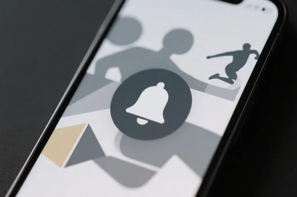 A close-up of a mobile phone screen displaying a stylized notification bell icon, surrounded by abstract geometric shapes representing sports motion, neutral and professional aesthetic